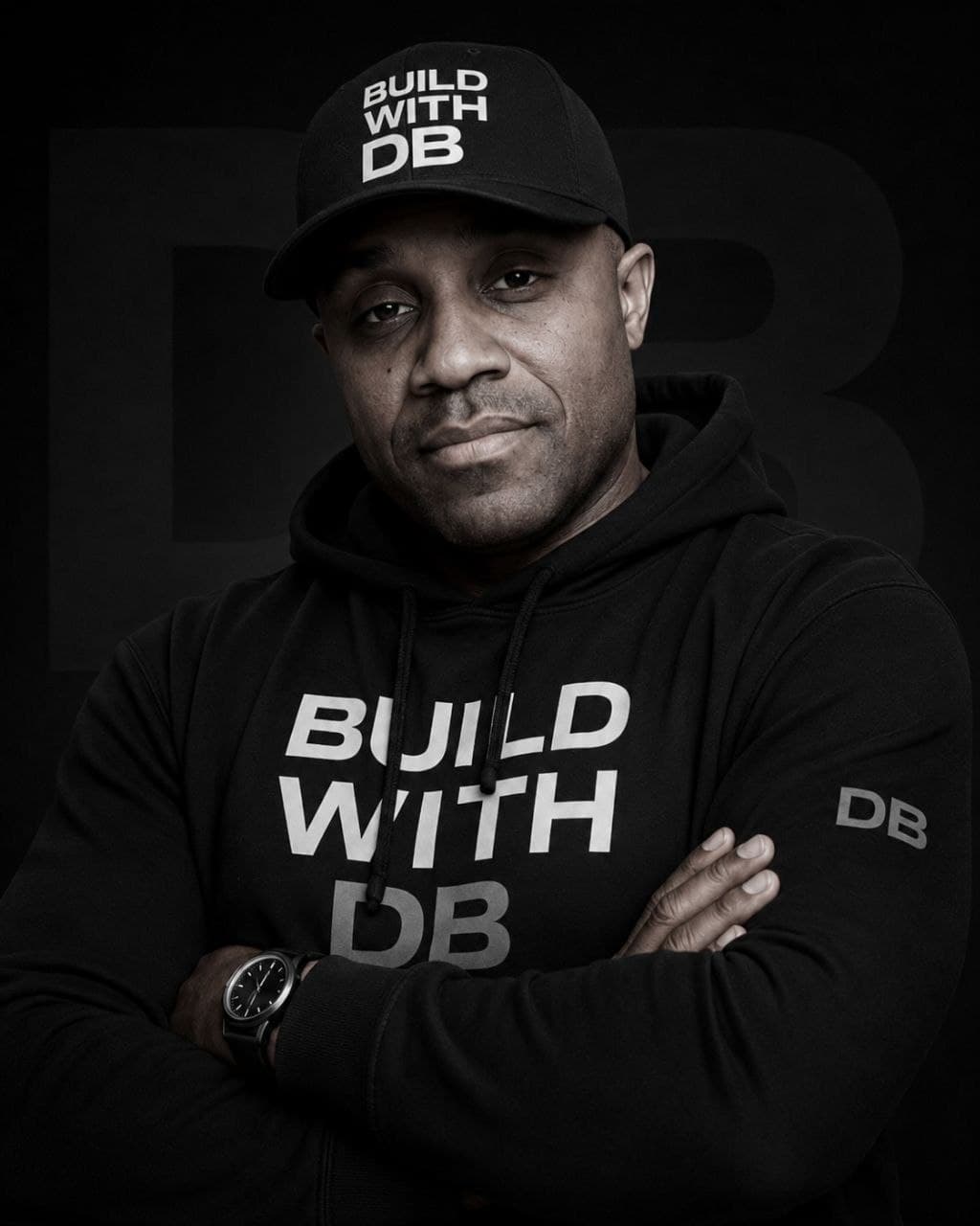 DB — arms crossed in a black Build With DB hoodie and cap, giant DB mark behind
